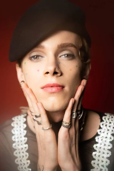 Portrait of nonbinary person in black beret and silver finger rings holding hands near face and looking at camera on dark red background — Photo de stock