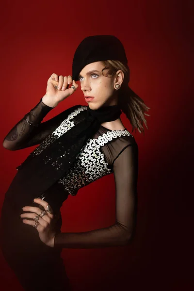 Queer person in elegant attire touching black beret while posing with hand on hip on dark red background — Foto stock