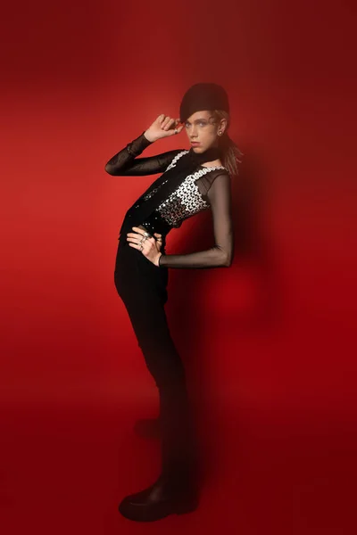 Toute la longueur du modèle queer à la mode ajustant béret noir tout en se tenant avec la main sur la hanche sur fond rouge foncé — Photo de stock