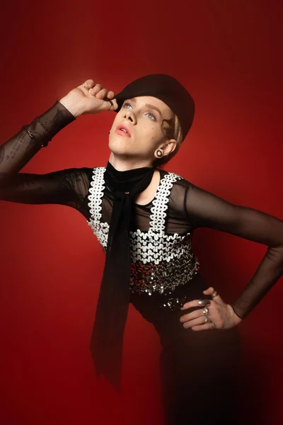 Fashionable nonbinary model holding hand on hip and touching black beret on red background - foto de stock