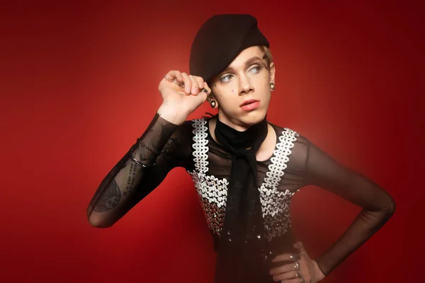 Fashionable queer model adjusting black beret while standing with hand on hip and looking away on red background - foto de stock