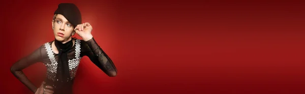 Young and stylish nonbinary person touching black beret and looking away on dark red background, banner — Fotografia de Stock