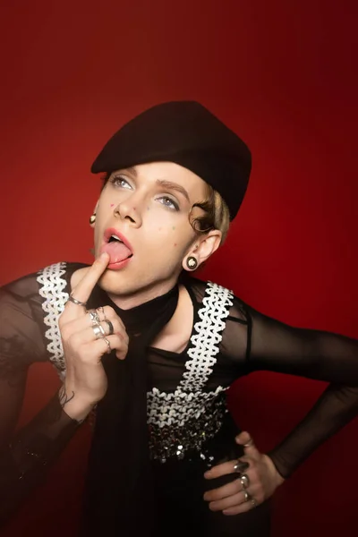 Nonbinary person in stylish outfit touching tongue with finger while looking away on red background — Stock Photo