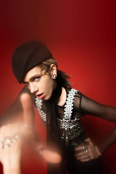 Stylish nonbinary model in black beret standing with hand on hip and pointing with blurred finger at camera on red background — Stockfoto