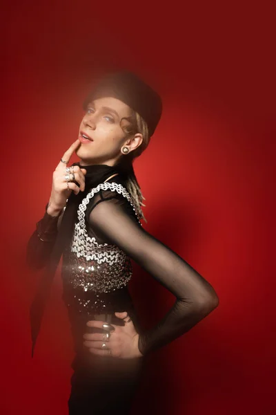 Elegant queer model in black beret touching lip while posing with hand on hip on red background — Photo de stock