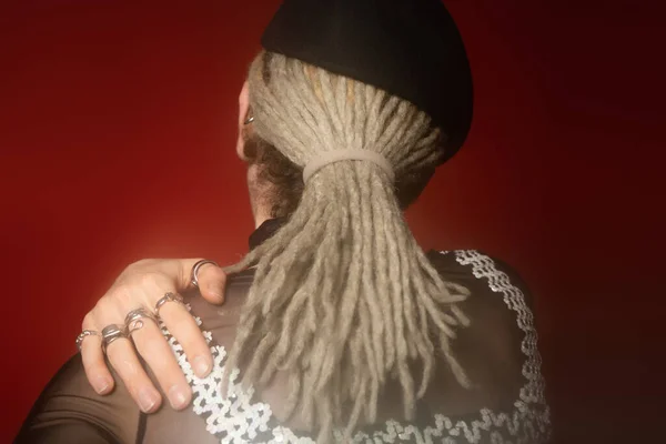 Back view of queer person with dreadlocks and silver rings holding hand on shoulder on dark red background — Stock Photo