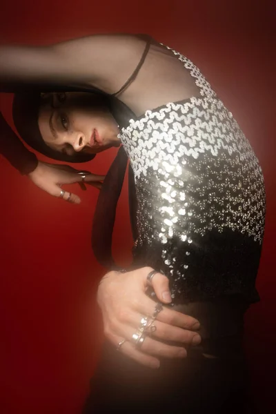 Glamour queer model in top with shiny sequins looking at camera while posing on red background - foto de stock