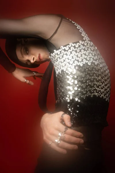 Glamour queer model in top with shiny sequins looking at camera while posing on red background — Stock Photo