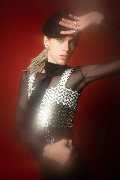 Trendy queer person in black beret and top with shiny sequins posing on dark red background — Stock Photo