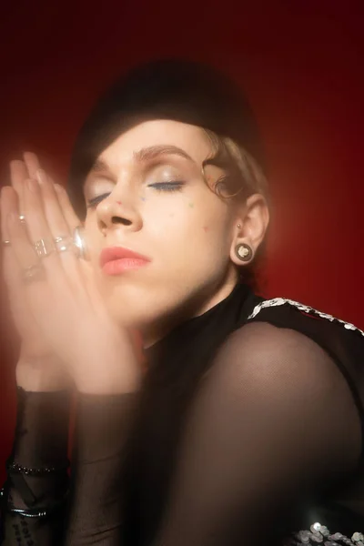 Portrait of blurred nonbinary person with closed eyes and praying hands on dark red background — Fotografia de Stock