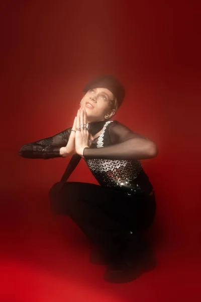 Full length of blurred queer person in trendy outfit posing with praying hands and looking up on dark red background - foto de stock