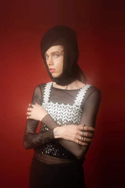 Nonbinary person in black headwear and top with sequins posing with crossed arms on dark red background — стоковое фото