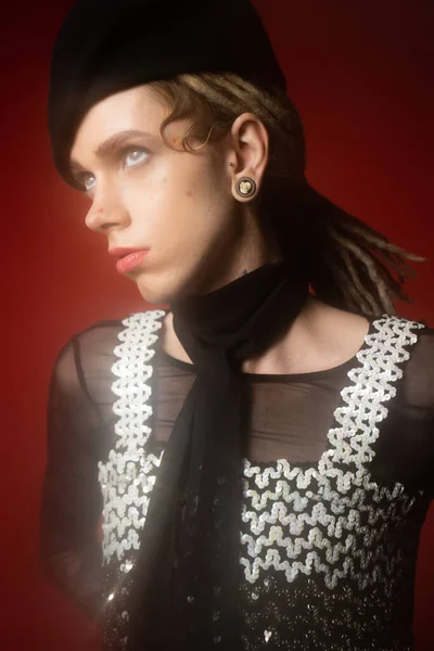 Trendy queer person in black scarf and top with sequins looking away on dark red background — стоковое фото