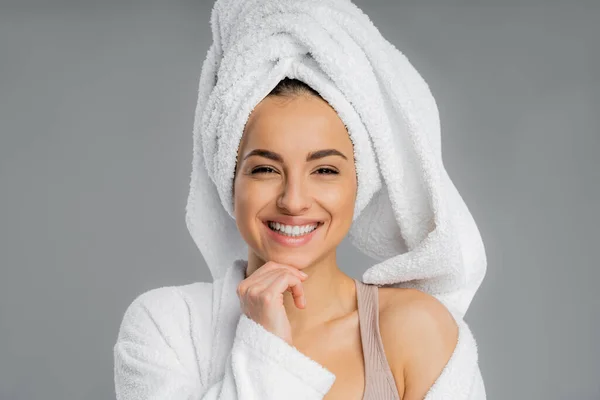 Cheerful young woman in bathrobe and towel on head isolated on grey — стоковое фото