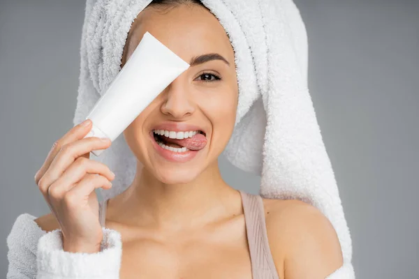 Young woman with towel on head sticking out tongue and holding cream isolated on grey — Photo de stock