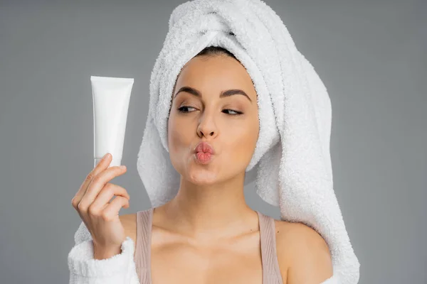 Young woman with towel on head holding cream and pouting lips isolated on grey — Stock Photo