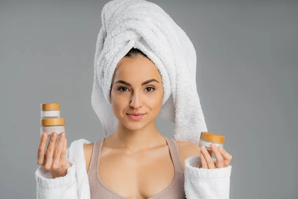 Young woman with towel on head holding creams isolated on grey — Photo de stock
