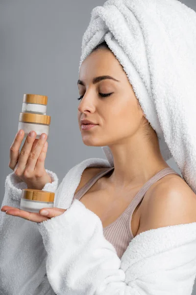 Woman in bathrobe and towel on head holding containers with creams isolated on grey — Photo de stock