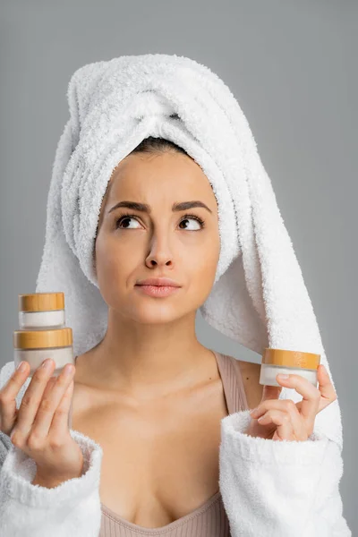 Pensive young woman with towel on head holding creams isolated on grey — Photo de stock