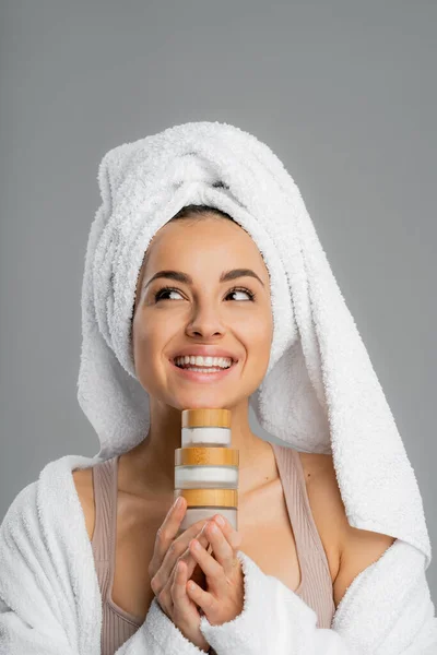 Positive young woman in towel on head holding cosmetic creams isolated on grey — Photo de stock