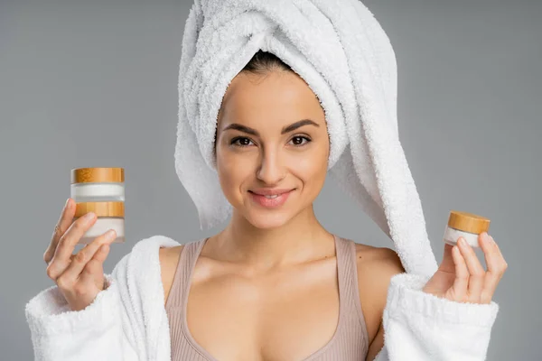Cheerful young woman with towel on head holding cosmetic creams isolated on grey — Stock Photo