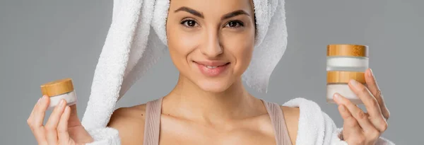 Smiling woman with towel on head holding cosmetic creams isolated on grey, banner — Photo de stock