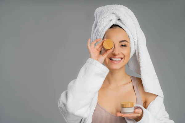 Smiling woman with towel on head holding cream near face isolated on grey — Stockfoto