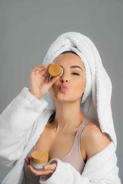 Woman in towel and bathrobe holding cosmetic cream and pouting lips isolated on grey — Stockfoto