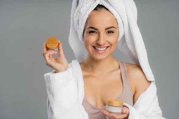 Cheerful young woman in towel holding cosmetic creams isolated on grey — Stockfoto