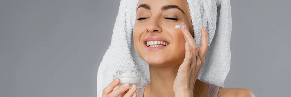 Cheerful young woman with towel on head applying face cream isolated on grey, banner — стоковое фото