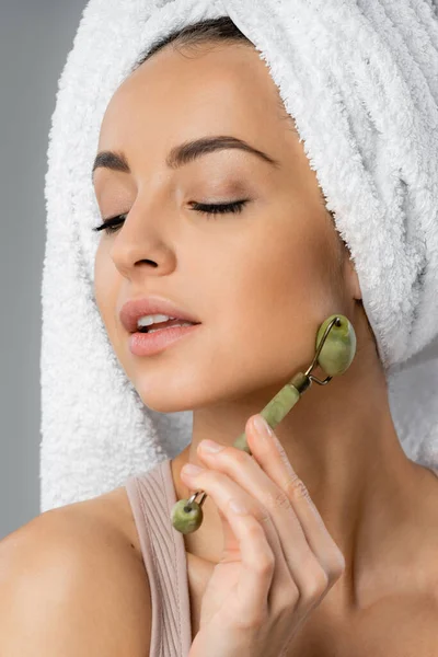 Portrait of young woman with towel on head using jade roller isolated on grey — Photo de stock