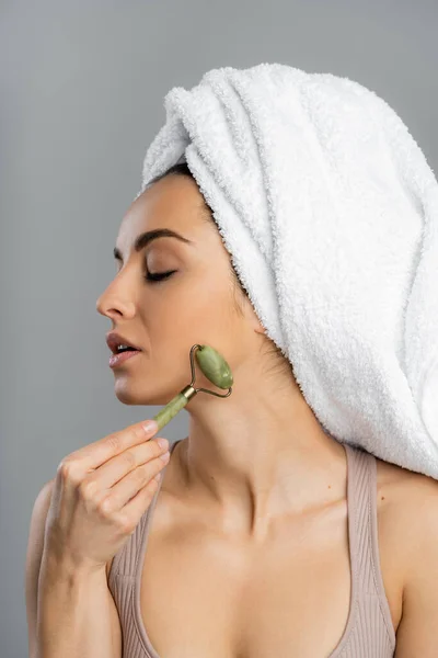 Pretty young woman with towel on head massaging face with jade roller isolated on grey — Fotografia de Stock