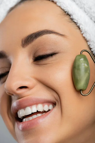 Close up view of smiling woman with towel on head massaging face with jade roller isolated on grey — Stock Photo