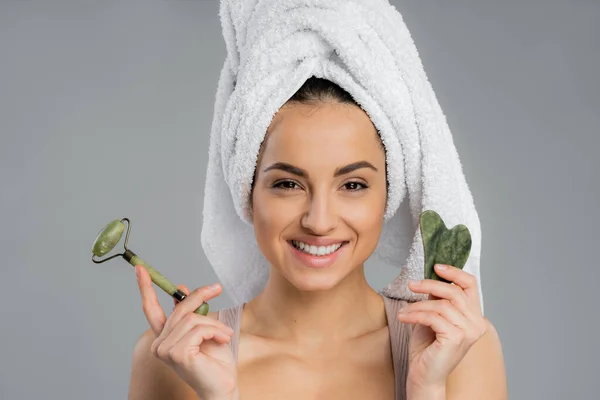 Smiling young woman with towel on head holding gua sha and jade roller isolated on grey — Fotografia de Stock