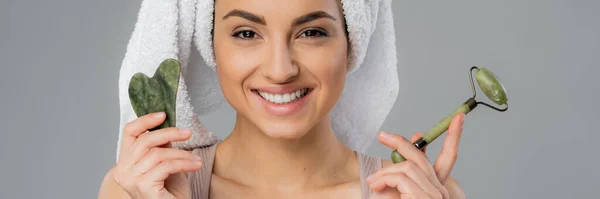 Cheerful woman with towel on head holding jade roller and gua sha isolated on grey, banner — Stockfoto
