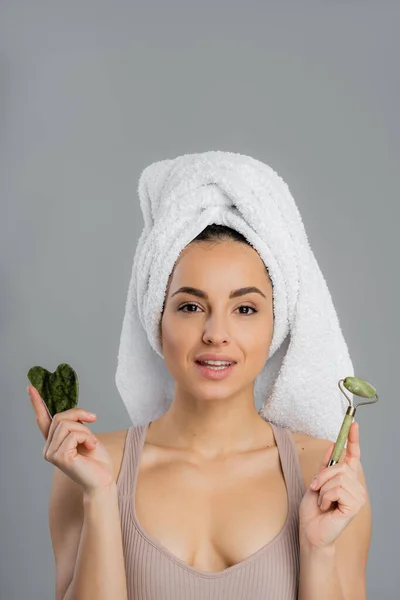Young woman with towel on head holding jade roller and gua sha isolated on grey — Fotografia de Stock