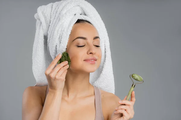 Pleased woman with towel on head using gua sha and holding jade roller isolated on grey — Stock Photo