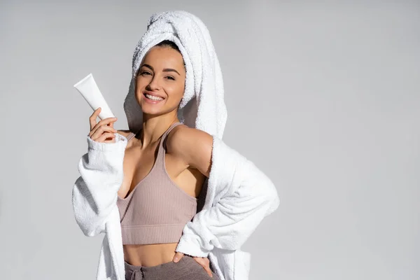 Cheerful young woman in towel and bathrobe holding cosmetic lotion isolated on grey — Foto stock