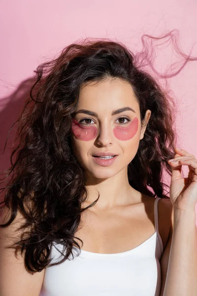 Curly young woman with hydrogel eye patches on pink background — стоковое фото