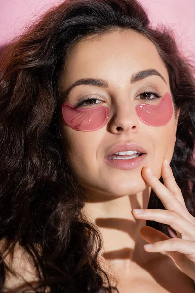 Portrait of curly woman with hydrogel eye patches looking at camera on pink background — Foto stock