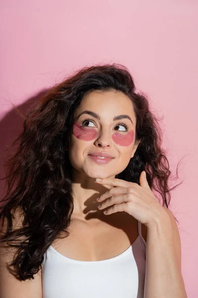 Dreamy young woman in top and hydrogel eye patches looking away on pink background — Stock Photo
