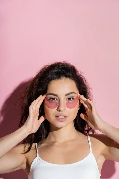 Pretty young woman in top touching hydrogel eye patches on pink background — Stock Photo