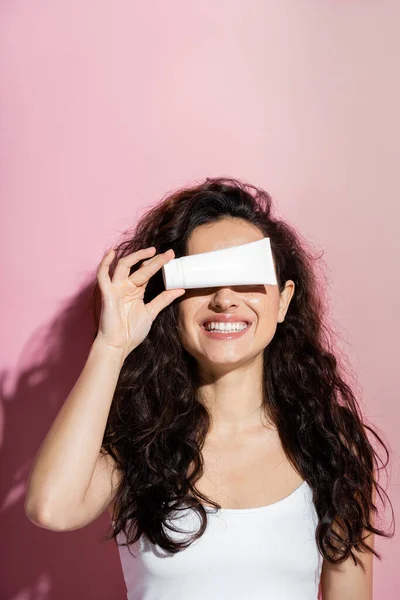 Smiling curly woman holding face cream on pink background — Stock Photo