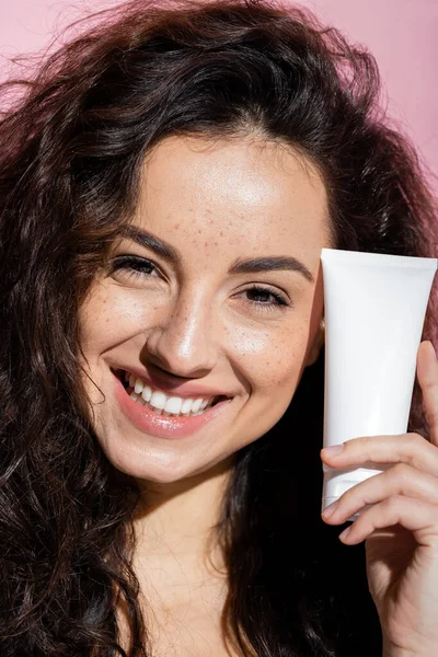 Cheerful young woman with freckles holding face cream isolated on pink — Stock Photo