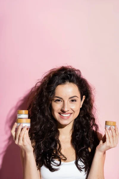 Curly young woman holding cosmetic creams on pink background — Photo de stock