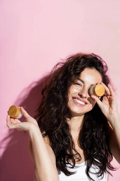 Positive curly woman holding jars with cosmetic creams on pink background — Stockfoto