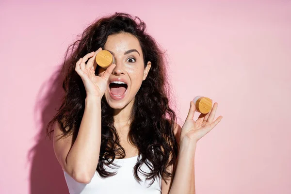 Shocked curly woman holding cosmetic creams on pink background — Stockfoto