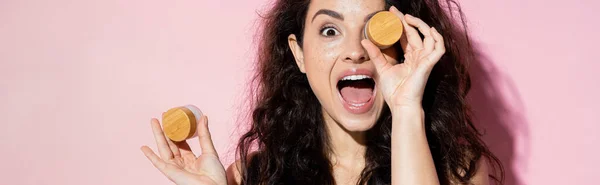 Shocked young woman holding jars with cosmetic creams on pink background, banner — Stock Photo
