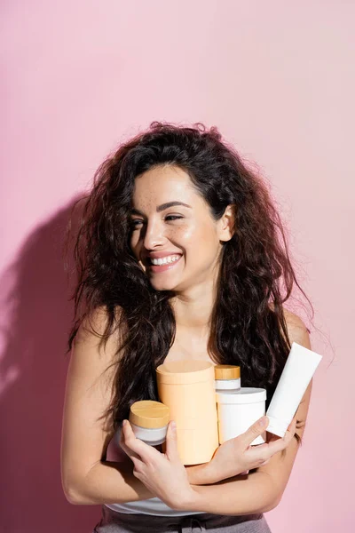 Cheerful curly woman holding cosmetic products on pink background — Photo de stock