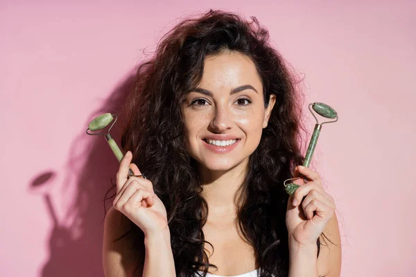 Cheerful freckled woman holding jade rollers on pink background — Fotografia de Stock